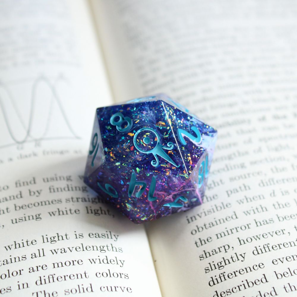A single jumbo d20 resting in-between the pages of a book. The die is blue and purple, and has lots of flecks of sparkling iridescence. The numbers are light blue.