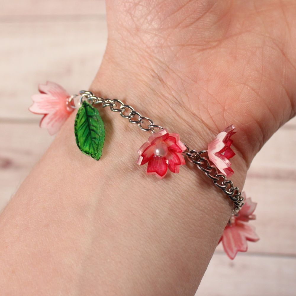 A bracelet made up of a silver chain with small pink flowers and green leaves attached to it, encircling a pale wrist.