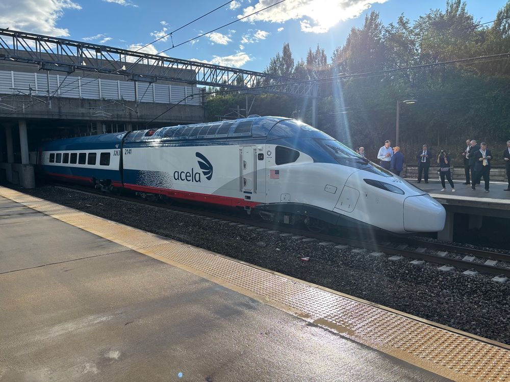 Avelia Liberty power car with striking pointed head end and vibrant blue and red accents and the Acela logo emblazoned prominently on the side emerges from under the Providence Amtrak under a bright blue late afternoon sky with sunlight flaring on the lens. People are gathered on the train platform behind the train.