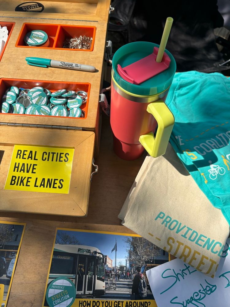 Close up of items arranged on a table under speckled sunlight. Promotional materials like postcards, teal pins and t shirts, canvas tote bags, and yellow sticker “REAL CITIES HAVE BIKE LANES” and at the center a tall colorful beverage container with a salmon pink body, teal lid, and yellow handle and straw.