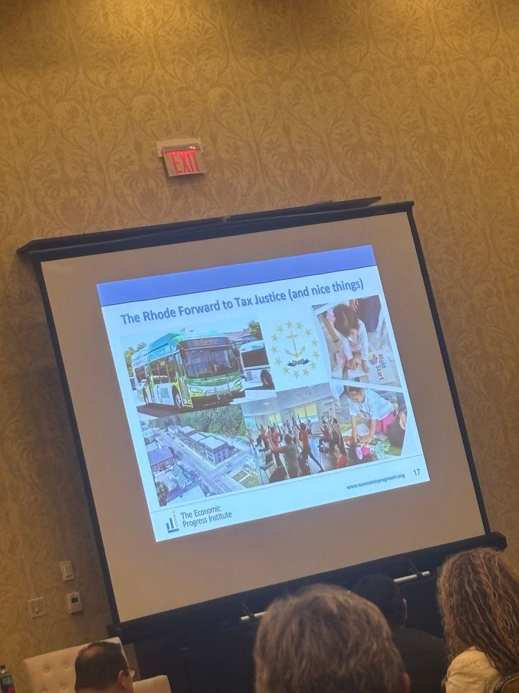 Presentation screen showing a slide with the title “The Rhode Forward to Tax Justice (and nice things)” showing multiple images including an electric Xcelsior RIPTA bus, the Rhode Island flag, small children playing, a yoga class, and an affordable housing development.