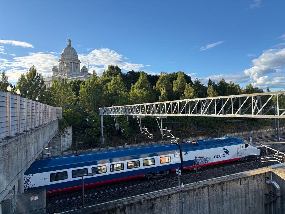 View of the Avelia Liberty trainset waiting in providence train station under the overhead gantry supporting the catenary wires with bright blue afternoon skies with some clouds visible. Behind a row of trees rising in the distance the dome of the Rhode Island State House is visible.