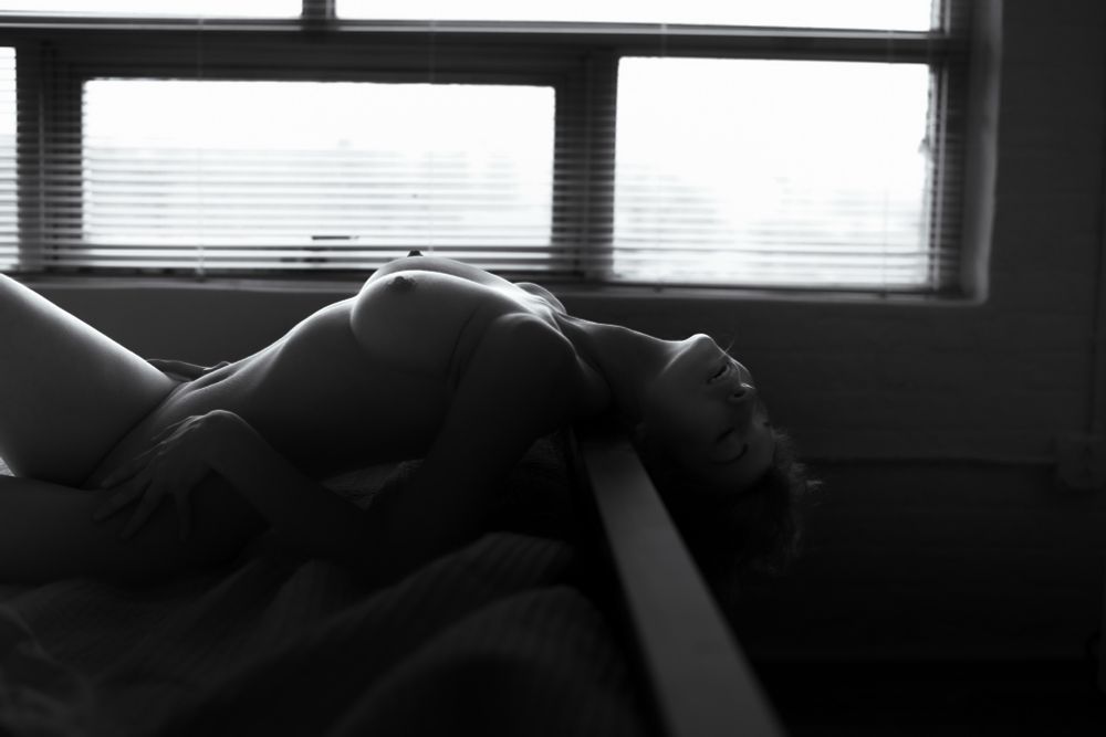 Backlit photo of a nude woman laying on a bed with her head hanging off the back of the footboard. 