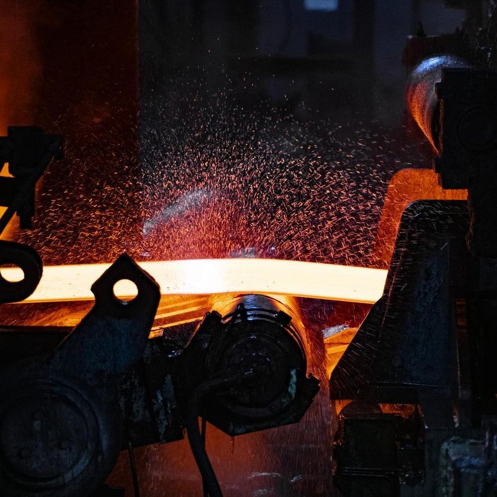 Ein glühend heißer Stahlbarren wird in einer dunklen Industrieumgebung durch Walzen gepresst.