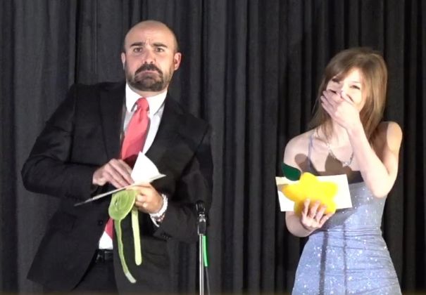 DataRich and KTWayfinder dressed on stage to present an award at the Kingdom Hearts Reconnect Million Dream Awards, Rich in a black suit with pink tie and KT in a blue dress. Rich has just ripped the envelope containing the winner in half and is looking directly at the camera with his brows furled and grimacing while KT covers her face to laugh