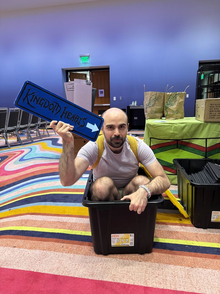 DataRich crouching in a black tote bin wearing a purple shirt and yellow backpack holding up a blue road sign that says “kingdom hearts” with an arrow pointed to himself
