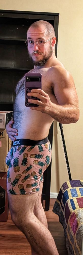 A hairy individual in pink underpants that have pineapples all over them. They're standing in profile, which accentuates both their butt and their package, filling out the underpants quite well.