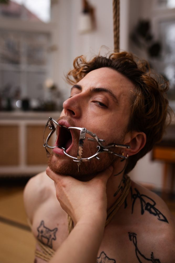 A man tied up in ropes bondage shibari / kinbaku style with a metal dental gag holding his mouth open.
The hand of the photographer is grabbing his neck, and he is glancing towards the viewer with half closed eyes.
He is blonde with black line tattoos on his neck and chest.