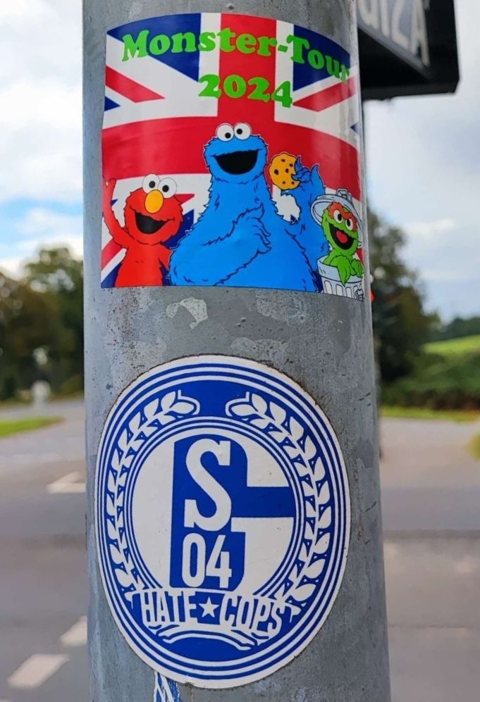 Ein Metallpfqhl eines Straßenschildes, auf dem ein SO4-Sticker und ein Sticker mit Sesamstraße-Monstern kleben.