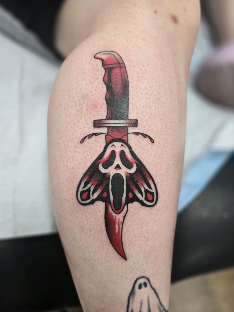 Picture of a tattoo on a leg. The tattoo is of a moth on top of a knife with the image of Ghostface worked into the moth's body and wings.