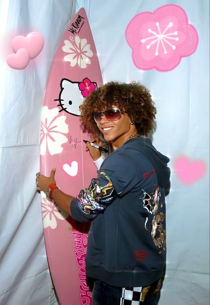 Corbin bleu de high school musical, ele é negro, tem o cabelo, grande, na altura do pescoço, crespo, loiro nas pontas, ele sorri pra tela, ele usa óculos de sol num tom caramelo, ele veste um moletom estampado da Nike, ele assina seu autógrafo numa prancha de surfe cor de rosa da Hello Kitty 