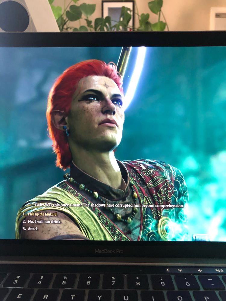 A picture of a MacBook running Baldur’s Gate 3. A male half-elf monk in dynamic blue-green lighting with red hair looks unamused at something off-screen. Dialogue text reads “Gale says: Was this once a man? The shadows have corrupted him far beyond comprehension.” Tav’s dialogue options read: One, action: pick up the tankard. Two, “No, I will not drink.” Three, action: ATTACK. 