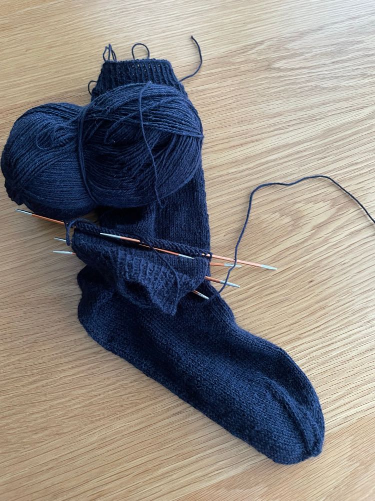Knitted sock and one in progress with cuff on needles. Dark navy. Wooden background 