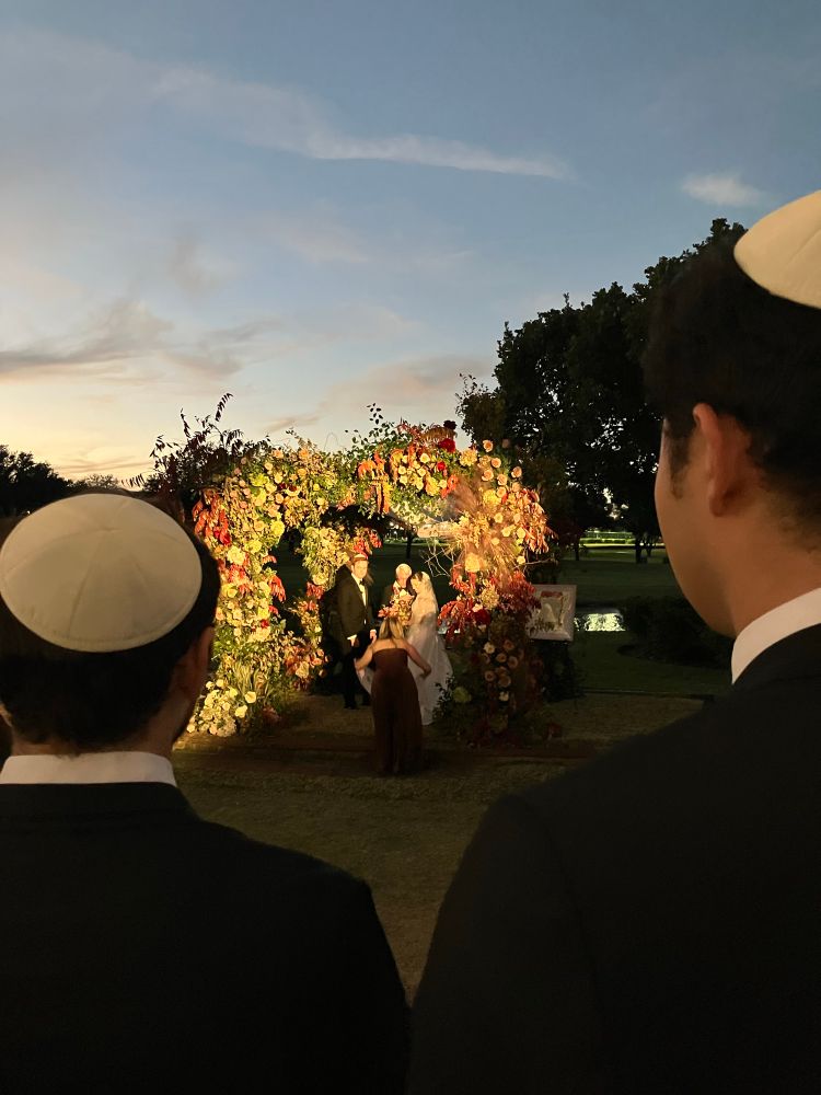 Bride and groom in a chuppa and two people in the foreground wearing yamakes sun setting in Texas golf course. Bridesmaids adjusting train. 
