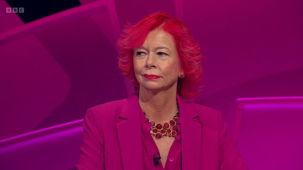 Pam, a woman wearing a fuschia pink blazer and matching blouse, statement necklace featuring large red stones set in gold with equally bright red hair and matching lipstick. The background is pink and she blends into it startling well.