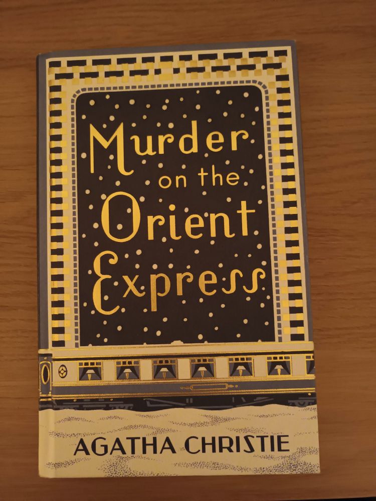 It's a beautiful gold, white and dark blue hardback of Agatha Christie's Murder on the Orient Express. It shows snow in the bottom quarter, topped by a first class dining carriage of a steam train and the remaining 2/3rd is dark blue sky with heavy snowflakes. There is a beautiful pattern around the edge. 