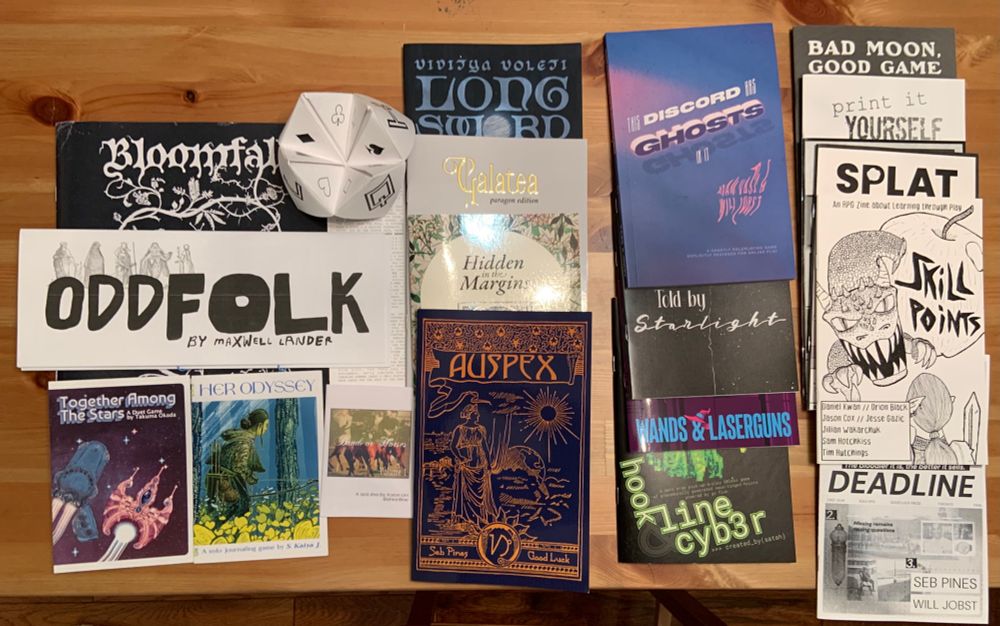 A collection of RPG zines arranged so the titles are visible. Included are:

Bloomfall by Sharang Biswas, Yeonsoo Julian Kim, and Joshua Gleason
Together Among the Stars by Takuma Okada
Galatea and Her Odyssey by S. Kaiya J.
Band of Horses by Aaron Lim
Oddfolk by Maxwell Lander
Slender Threads of Freqs and Fortune and Hook, Line & Cyber by Satah (Wicked Glitch Games)
Hidden in the Margins and Deadline by Will Jobst and Seb Pines (Good Luck Press)
Auspex by Seb Pines
This Discord Has Ghosts in It by Adam Vass and Will Jobst
Longsword by Viditya Voleti
Told by Starlight by Ryan Khan
Wands & Laserguns by Christian Bahnweg and Justin Vandermeer
Bad Moon, Good Game and Print it Yourself by Justin Vandermeer (Shouting Crow)
SPLAT 3, SPLAT 4, and Skill Points by Summoning Circle Press. 