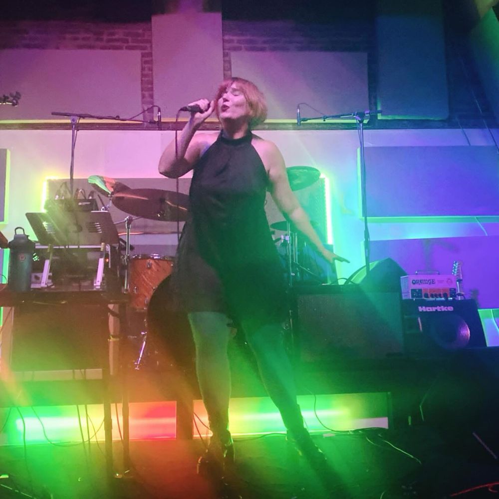 A colorfully lit stage with me singing. I’m wearing a black satin halter romper, green tights, black boots. 