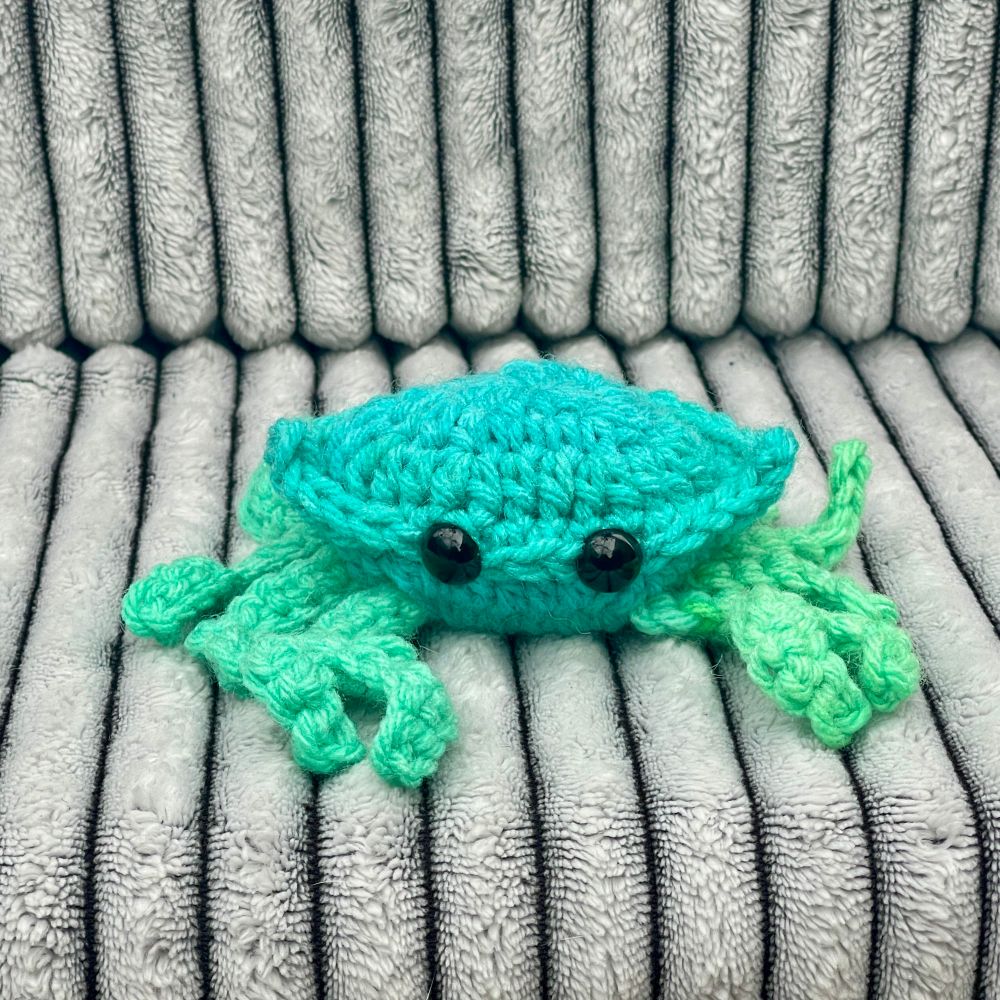 A blue-green crocheted crab with beady black eyes.