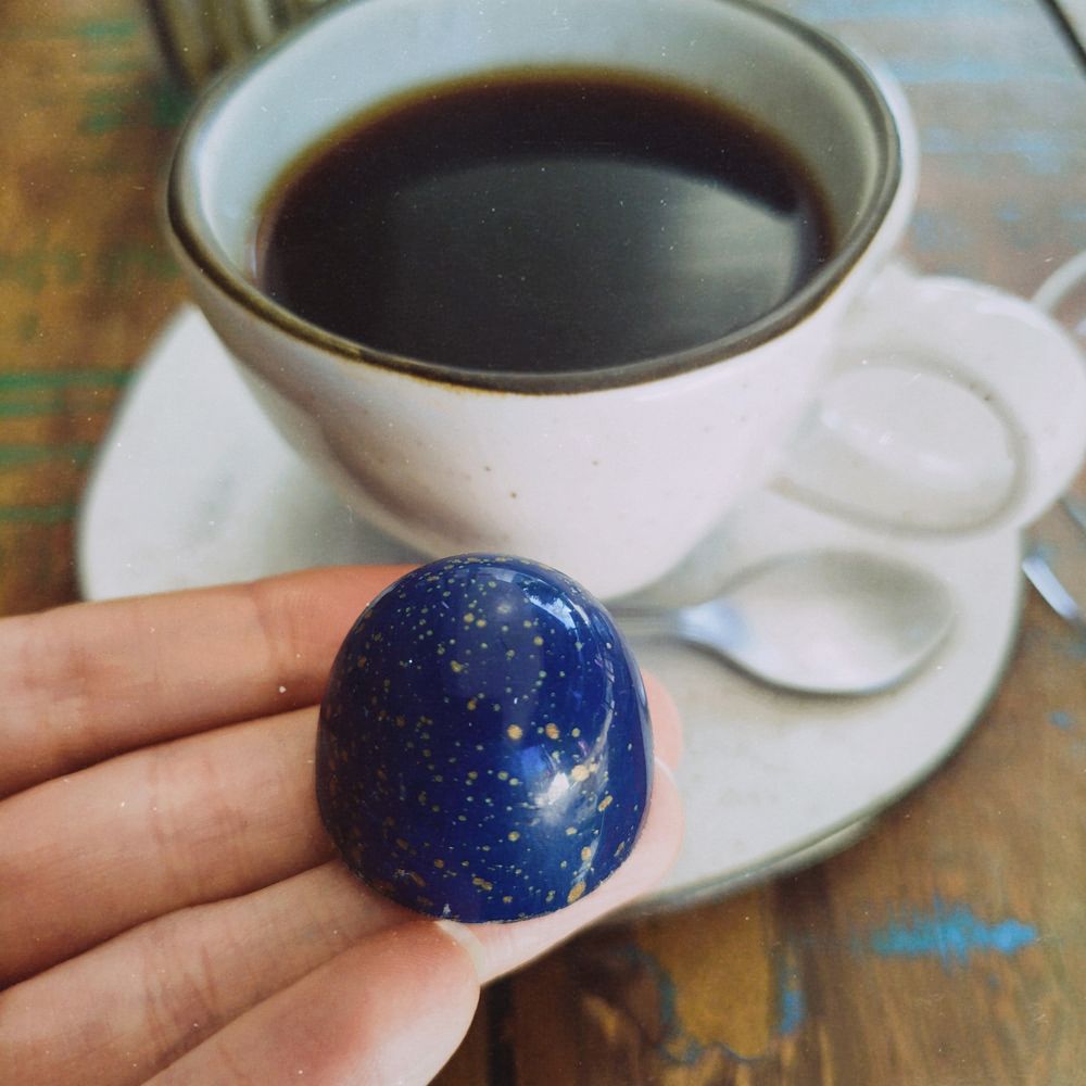 Picture of my hand in close up holding a chocolate bon-bon painted in dark blue with golden speckles, with a black coffee cup on the background.