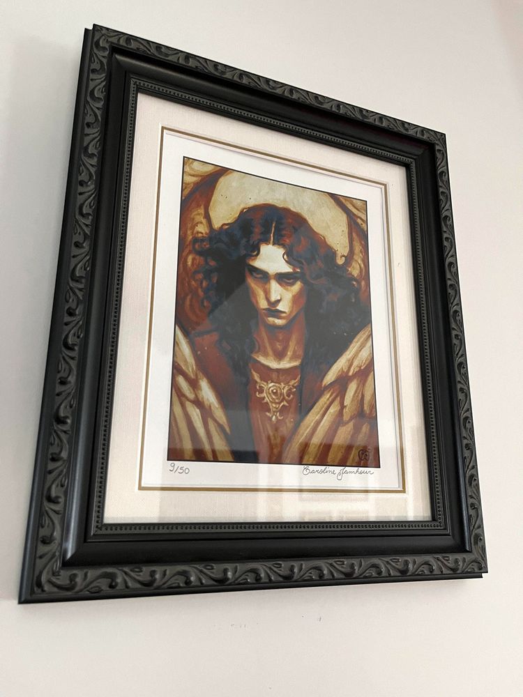 Picture of a print of my artwork "Shadowed Grace", which depicts a stern looking angel in muted orange tones, in a black ornamented frame.