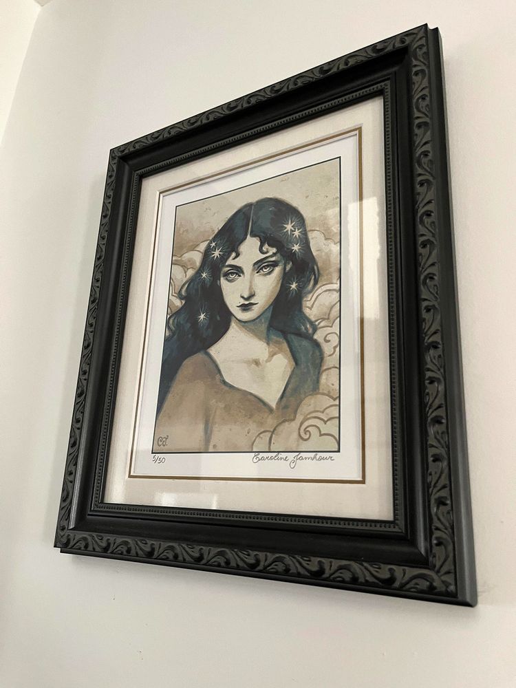 Picture of a print of my artwork "Celestial Muse", which depicts a woman among clouds with stars on her hair, in a black ornamented frame.