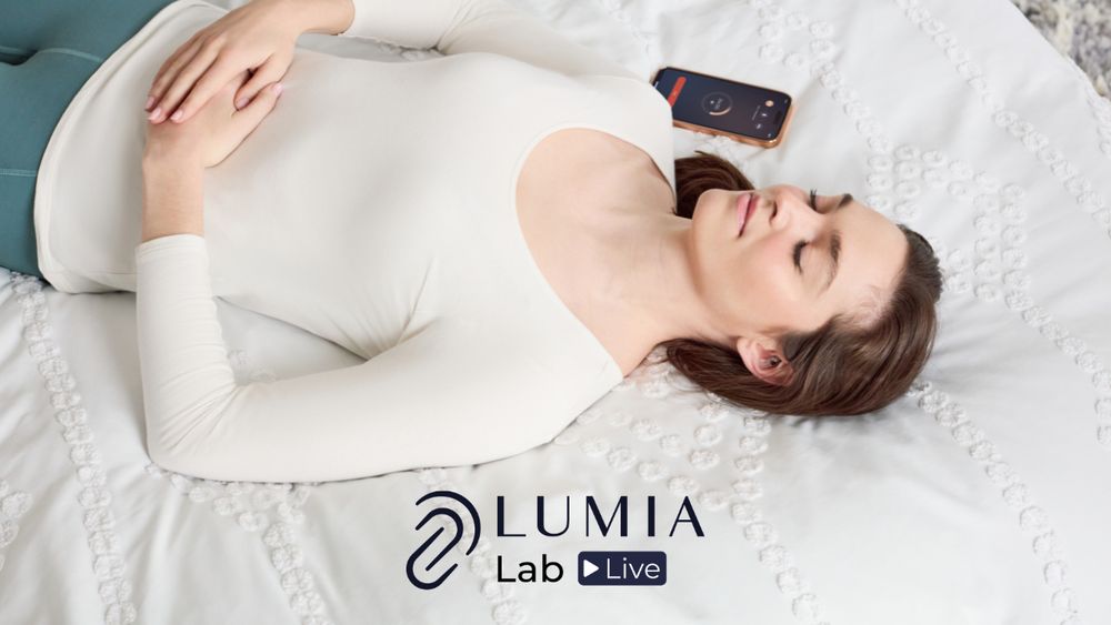 Text reads Lumia Lab Live. A caucasian female with hypermobile EDS, POTS and Long Covid Dysautonomia rests on a white bed with her eyes closed, a Lumia wearable device is visible in her ear on close inspection. An iPhone is lying next to her running Live Mode data collection on the blood flow to her head as well as heart rate.