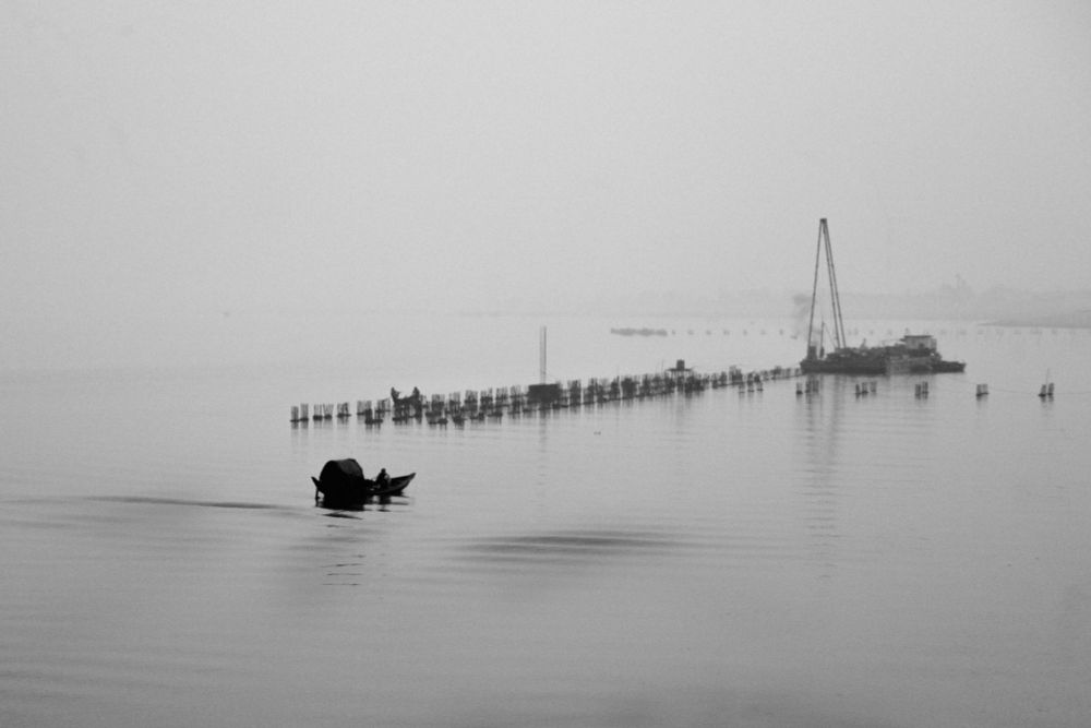 A black and white photo of a boat reaching towards a hazy line of trees that might lead to an island which is the boatman's home