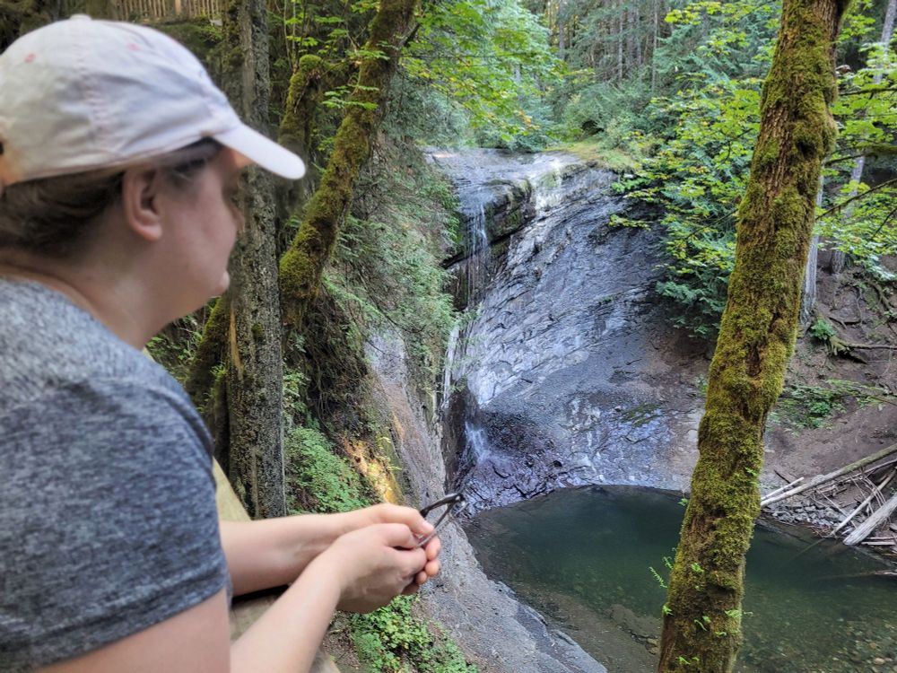 A person in a white hat and a grey shirt looking down through moss-covered trees at a waterfall and pool, Ammonite Falls, BC, Canada.