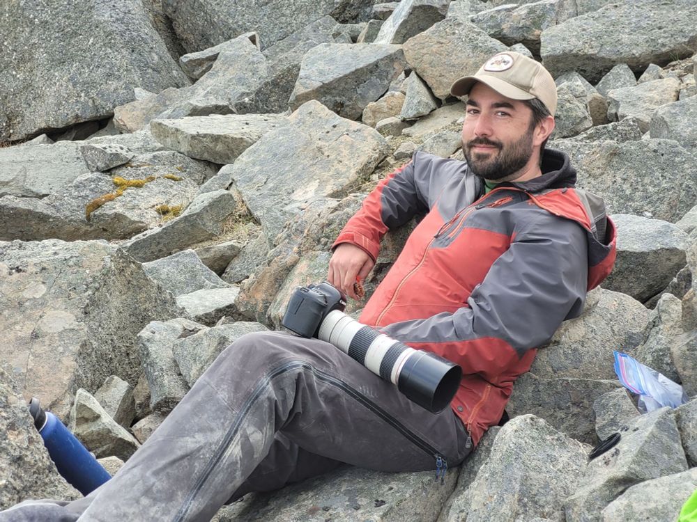 Dr. Thomas Cullen, in a red jacket with camera in his lap, reclines on the grey rocks of Spatsizi Plateau.