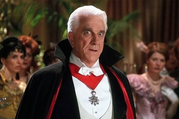 Leslie Nielsen (homem branco, grisalho) vestido de Conde Drácula, com uma capa preta com forro vermelho, camisa branca e detalhe vermelho com ponta de metal no pescoço. Cena do filme Drácula morto mas feliz.