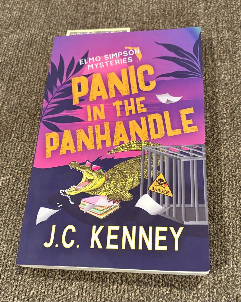 Book title: Panic in the Panhandle (Elmo Simpson Mysteries)
Author: J.C. Kenney 
Book description: The book is on my sofa so the cover shows clearly. The cover is a pink/purple on the top half showing the title in gold. On the bottom half of the cover there is an alligator wearing pink sunglasses with an open jaw. There are pearls in its mouth and its left front paw is on top of colorful spiral-bound notebooks. Papers are strewn about. And there’s a cage with a danger (skull and crossbones) symbol on it with what looks like blood smeared on the danger sign, too.
