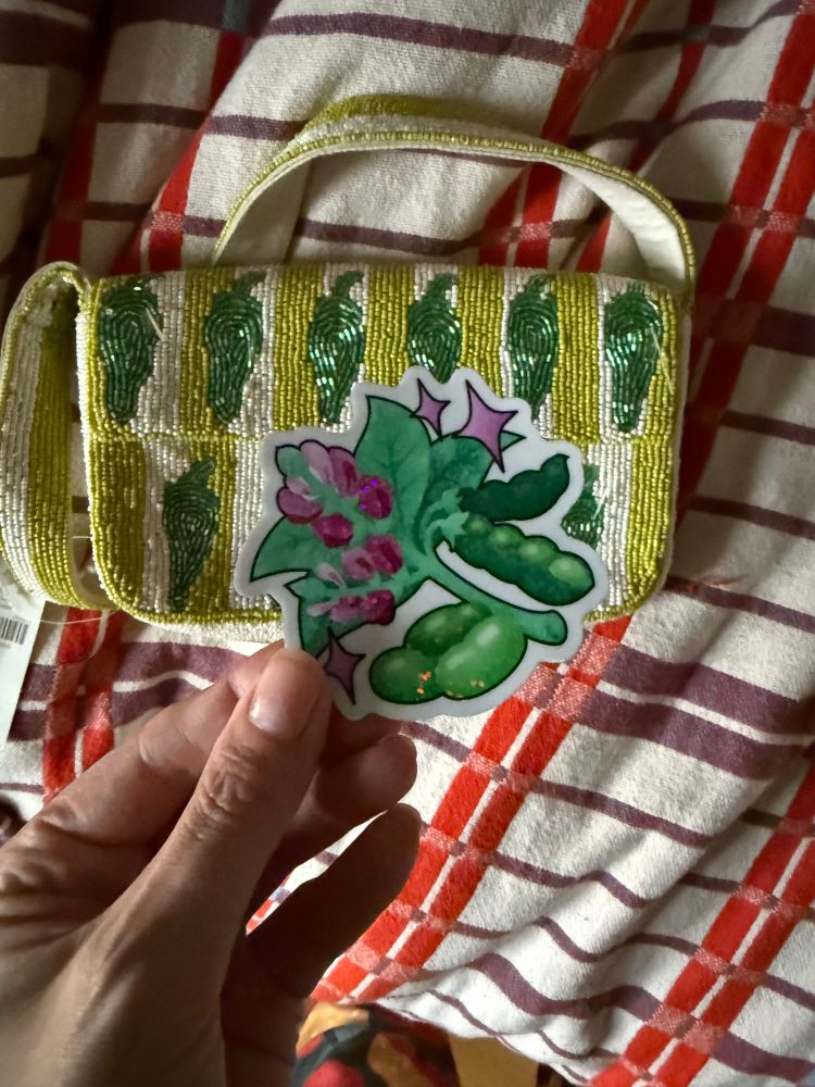 A chartreuse and cream striped beaded purse with darker green peppers on it on a red plaid coverlet, a white hand is holding holographic bean sticker 