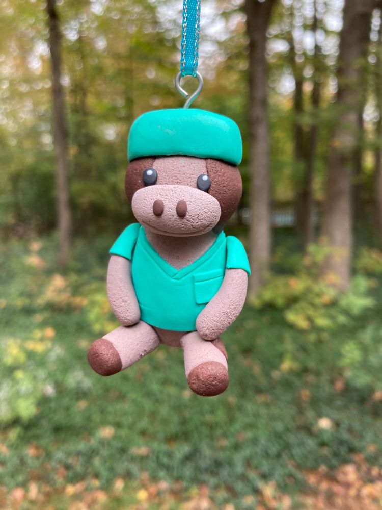 Clay ornament of a tan and brown cow stuffie, dressed in a green scrub shirt and green scrub cap. The ornament hangs from a teal and silver ribbon in front of a forest.