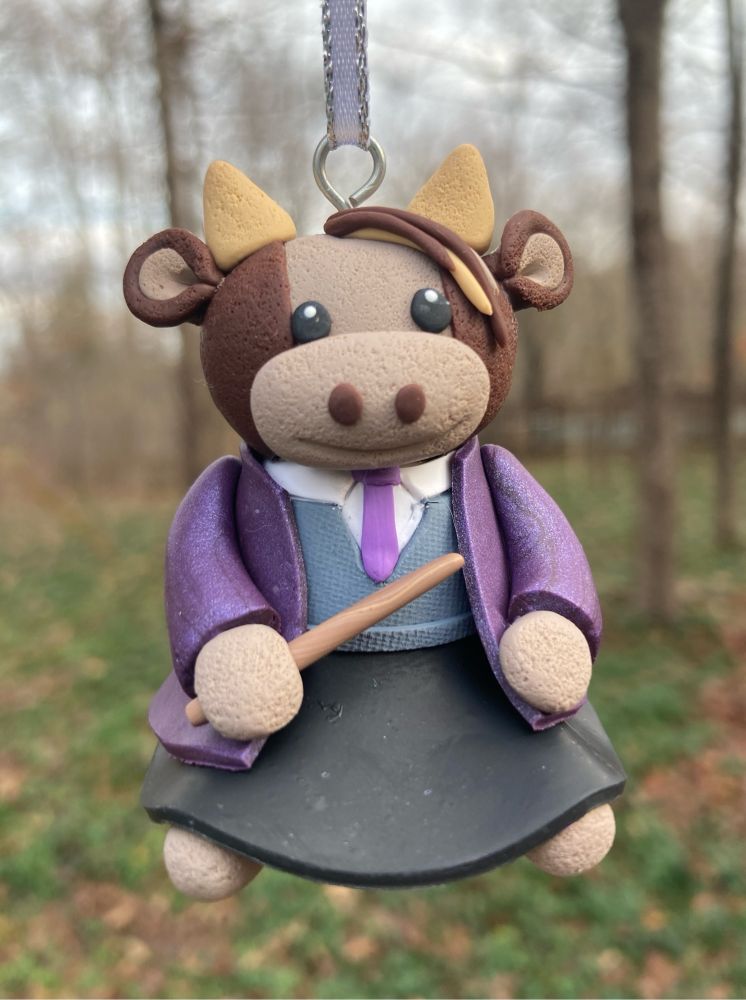Clay ornament of a tan and brown cow stuffie, wearing a preppy school uniform- black skirt, white collared shirt, grey vest, and purple tie -under a sparkly purple long-sleeved robe. It holds a smallmagic wand. The ornament hangs from a white and silver ribbon in front of a forest.