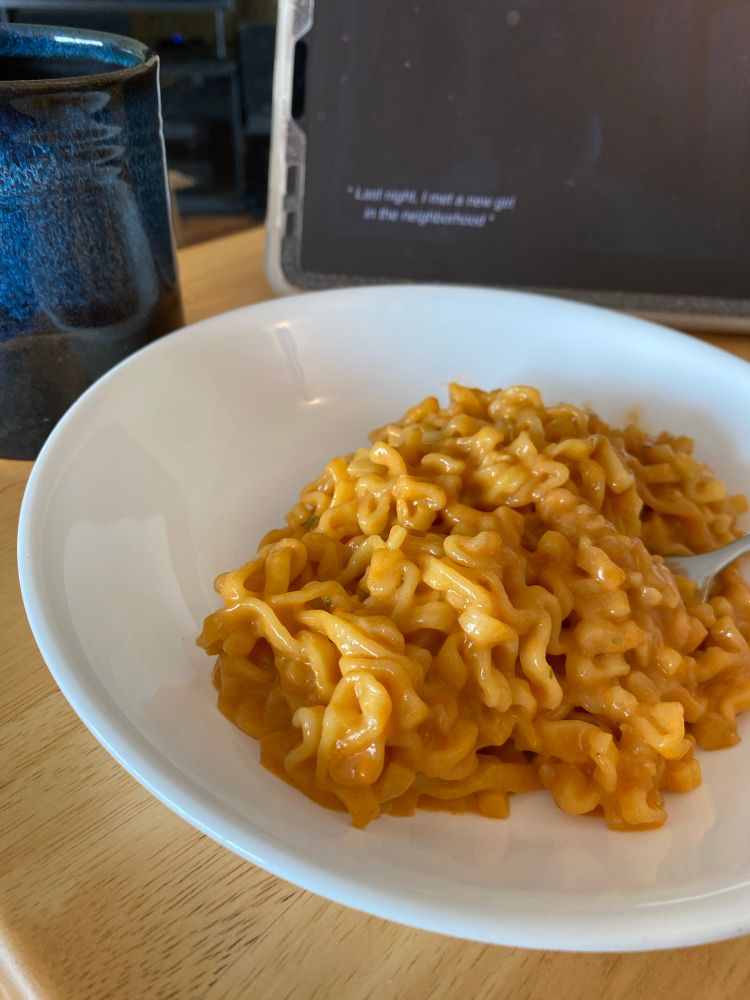 An image of spicy noodles on a plate with a mug of coffee and a corner of an iPad with Hunting Party playing in the background.