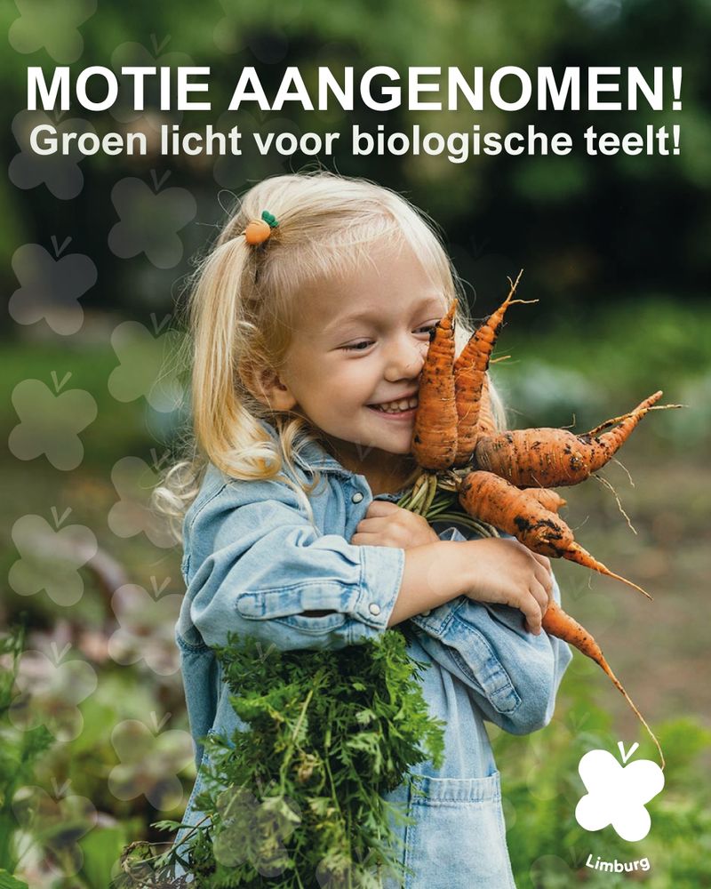 Een blond jong meisje in een lichtblauwe spijkerjurk lacht vrolijk. In haar armen heeft ze een bos vers geplukte wortelen en houdt deze tegen haar gezicht aan. De tekst boven de foto is: Motie aangenomen! Groen licht voor biologische teelt!