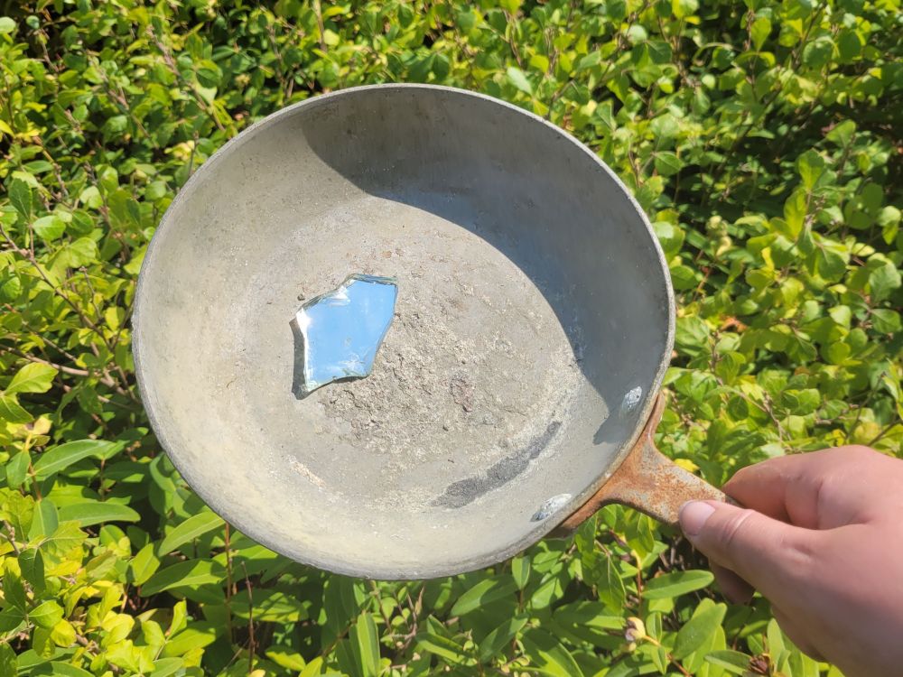 my hand in lower right corner holding a small frypan found at lowtide with a bit of mirror (found on trail) inside. its blue from the sky. the frypan is all dry silt and rust