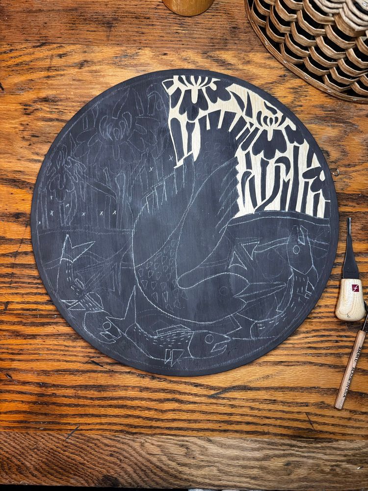 A partially carved wood round of a duck peering under the water looking at fish. Behind the duck you can see irises growing along the waters edge. 