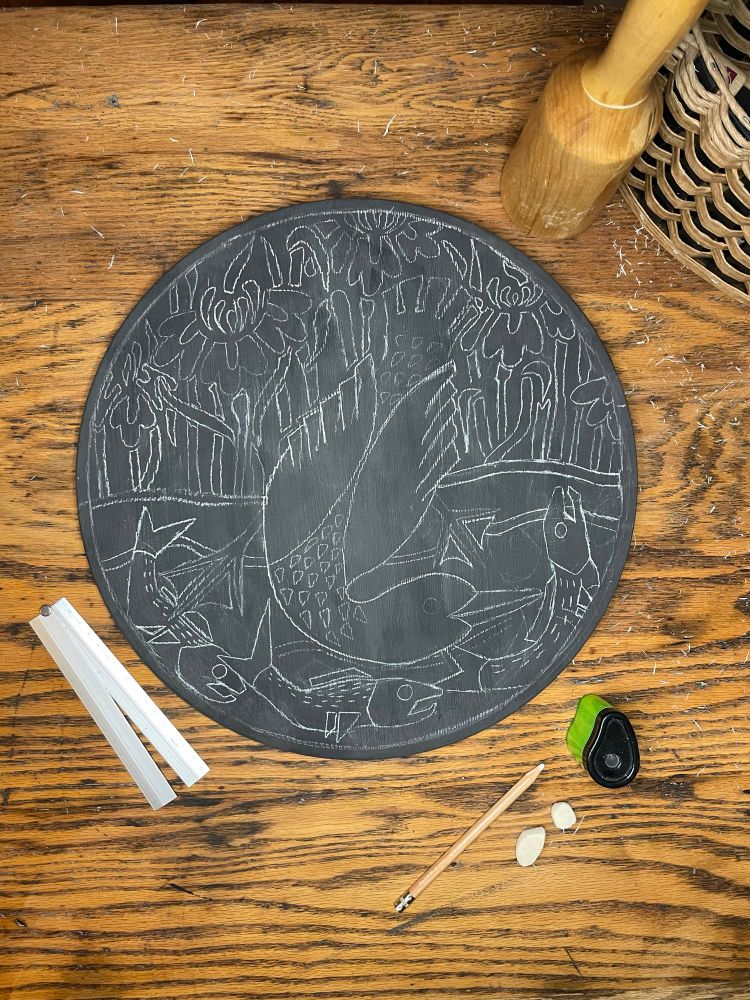 Top down view onto a desk where a wood round with a chalk drawing sits waiting to be carved. Surrounded by the tools needed to draw the piece, chalk pencil, pencil sharpener, erasers, etc. 
