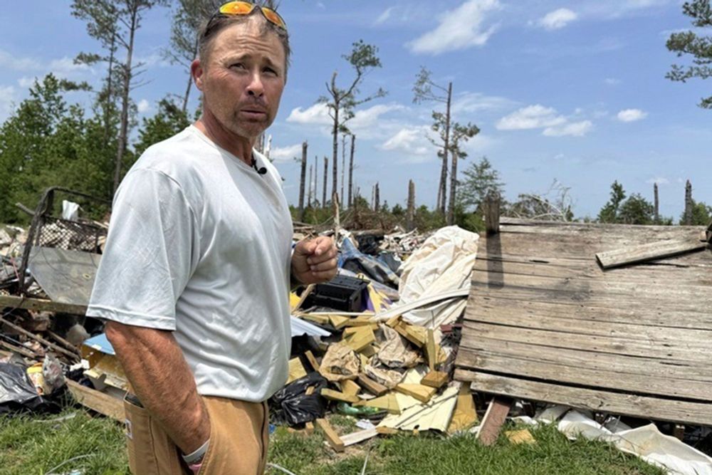 Man in grey shirt and khaki's stand in front of a pile of debris in an open field 
