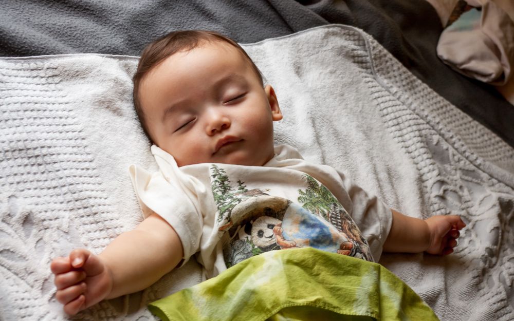 In this color photograph a baby is sleeping on his back with his arms spread wide like the baby Jesus. The natural light from a window adds a softness to the image.