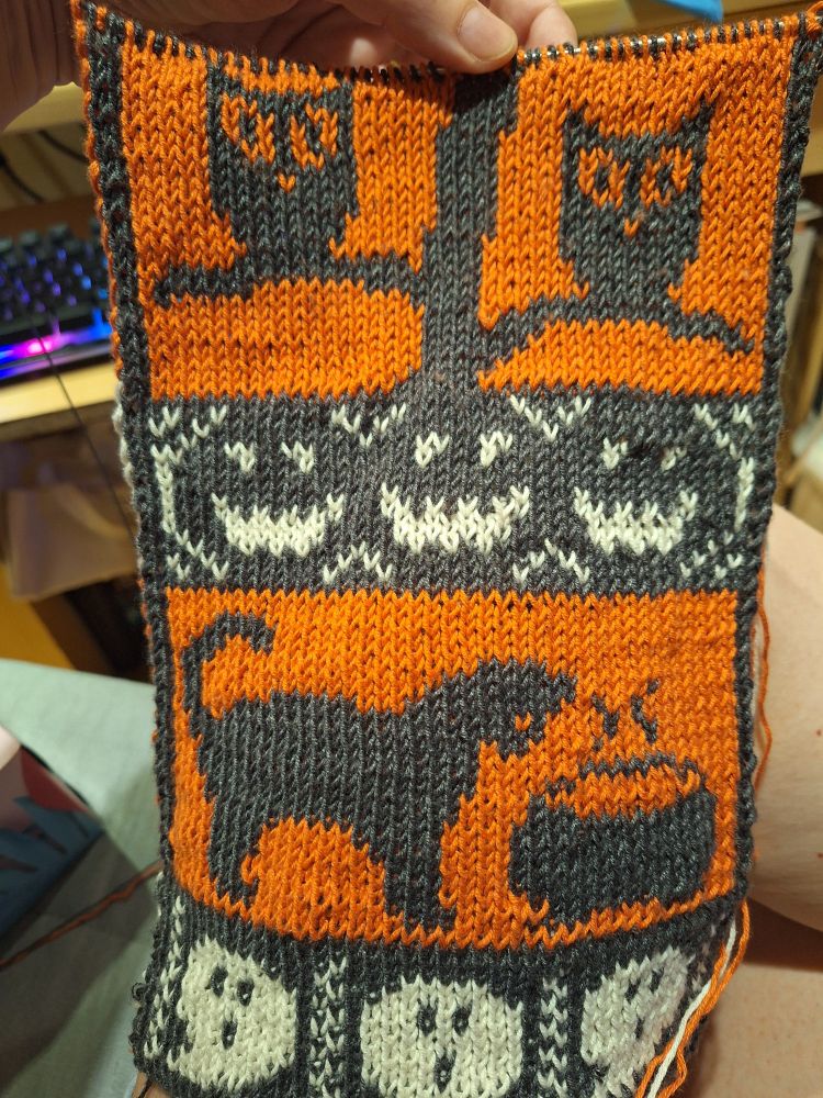 Photo of a scarf being knit. A hand holds up the beginnings of a Halloween themed scarf. It's made with three colours: orange, white and dark grey. It has small panels depicting different scenes on top of one another. From the bottom up there are ghosts, a black cat looking at a cauldron, three jack-o-lanterns and two owls sitting on a tree.