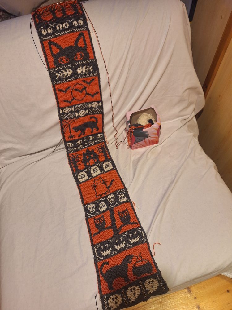 Photo of a scarf draped on a couch. The scarf is still being knit. The scarf depicts black-orange-white panels of small spooky scenes with bats, spiders, ghosts, jack-o-lanterns, candy and lots of black cats.
