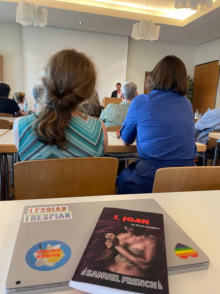 A laptop and the playtext of I, Joan on a table, people seated on chairs listening to Charlie Josephine, who is in the back, speaking at a podium. 