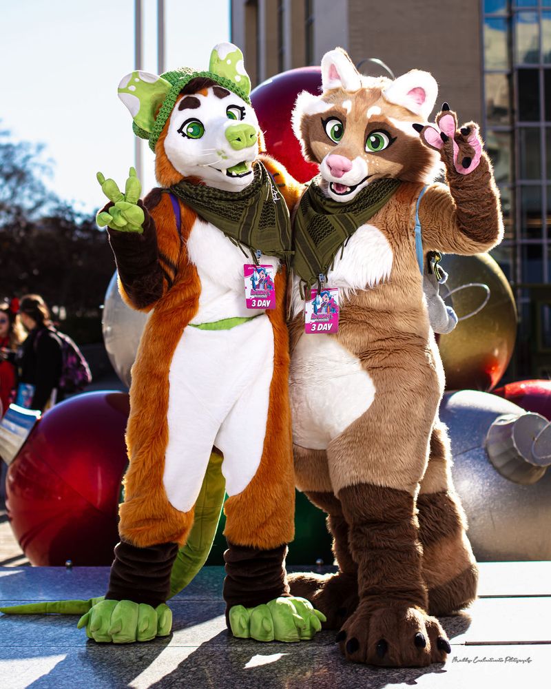 Photo of two fursuiters posing together. There’s an opossum with green accents on the left, and a cinnamon raccoon with a pink nose on the right. They are both wearing matching green bandanas, and the opossum also has a knitted green beanie. 