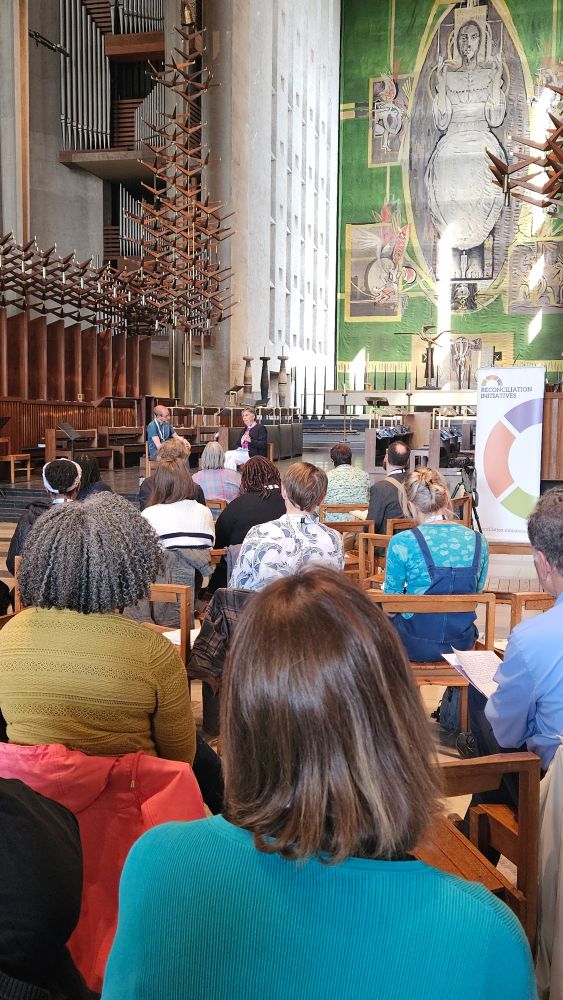 People sat in rows at Coventry cathedral. 2 speakers at the front. Wall hanging in background of Jesus 
