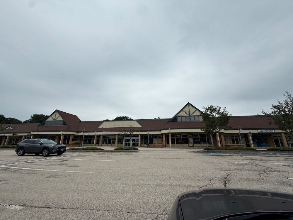 Sad, empty shopping plaza, foregrounded by mostly empty parking lot, under gloomy skies. 