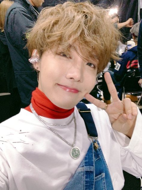 J-hope, wearing denim overalls and a white sweatshirt over a red turtleneck, smiling and making a V-sign at the camera.