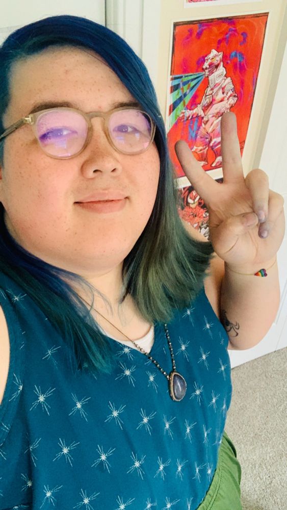 Zoey Reyes, a light skinned asian woman with blue hair just past shoulder length, wearing a blue patterned sleeveless top, a green skirt, glasses, and a necklace with an embedded blue stone in a teardrop shape.